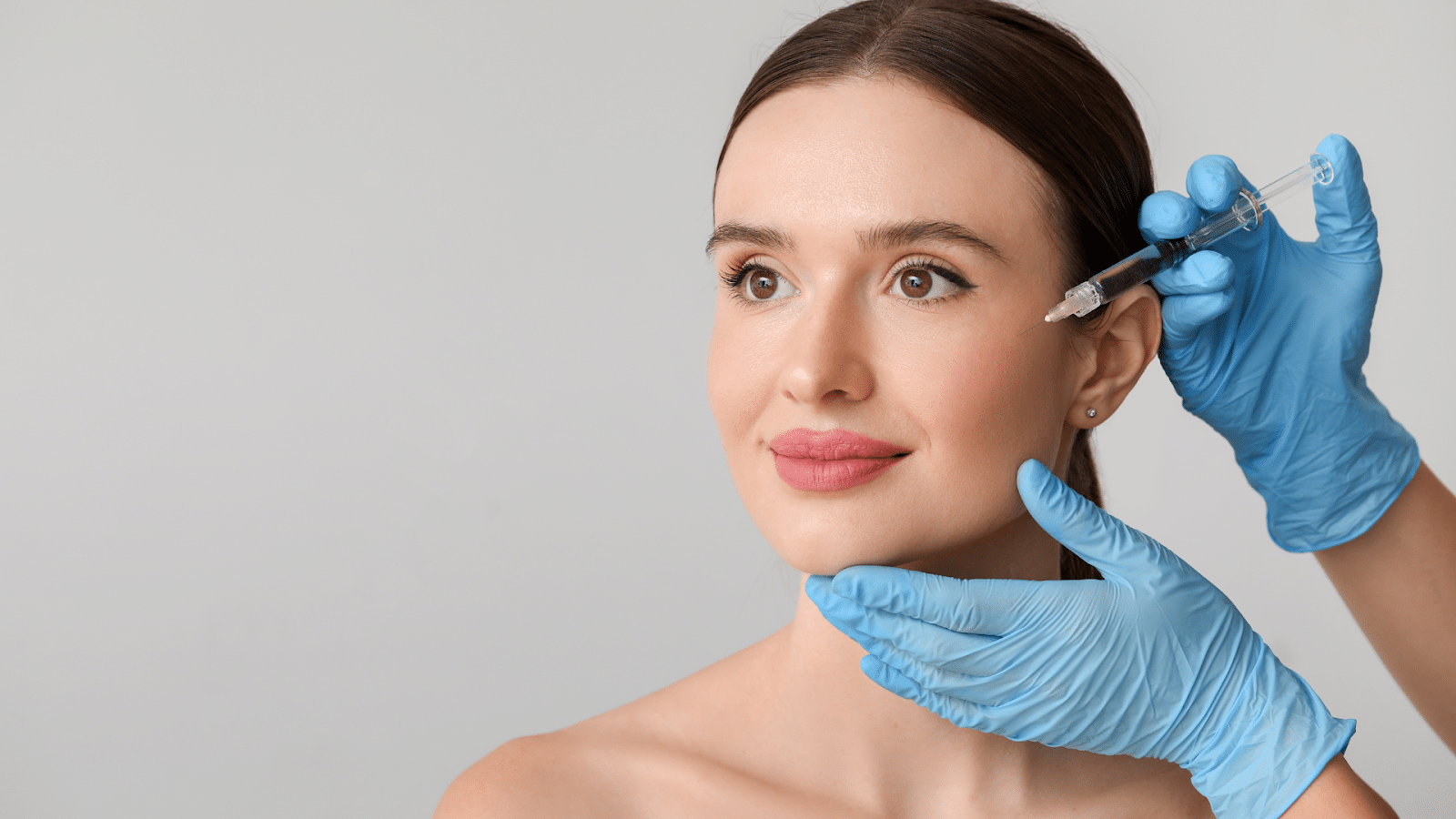 A woman with a serene expression receives an injection in her cheek from someone wearing blue gloves. The background is a simple gray, and the woman's hair is neatly pulled back.