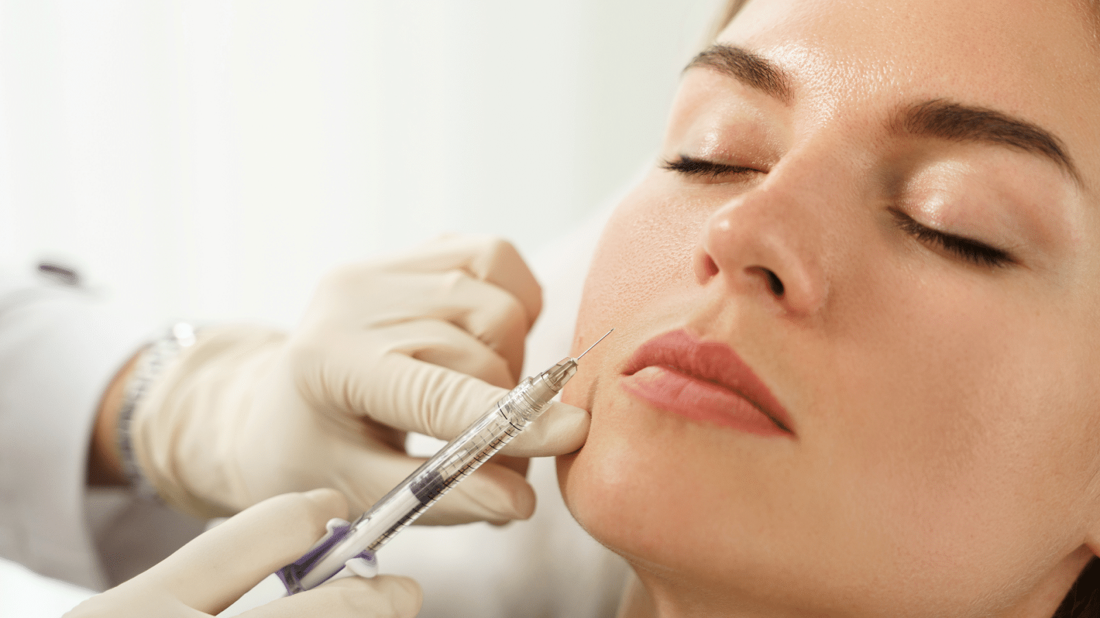 A person with closed eyes receives a cosmetic injection in the cheek from a gloved medical professional using a syringe.