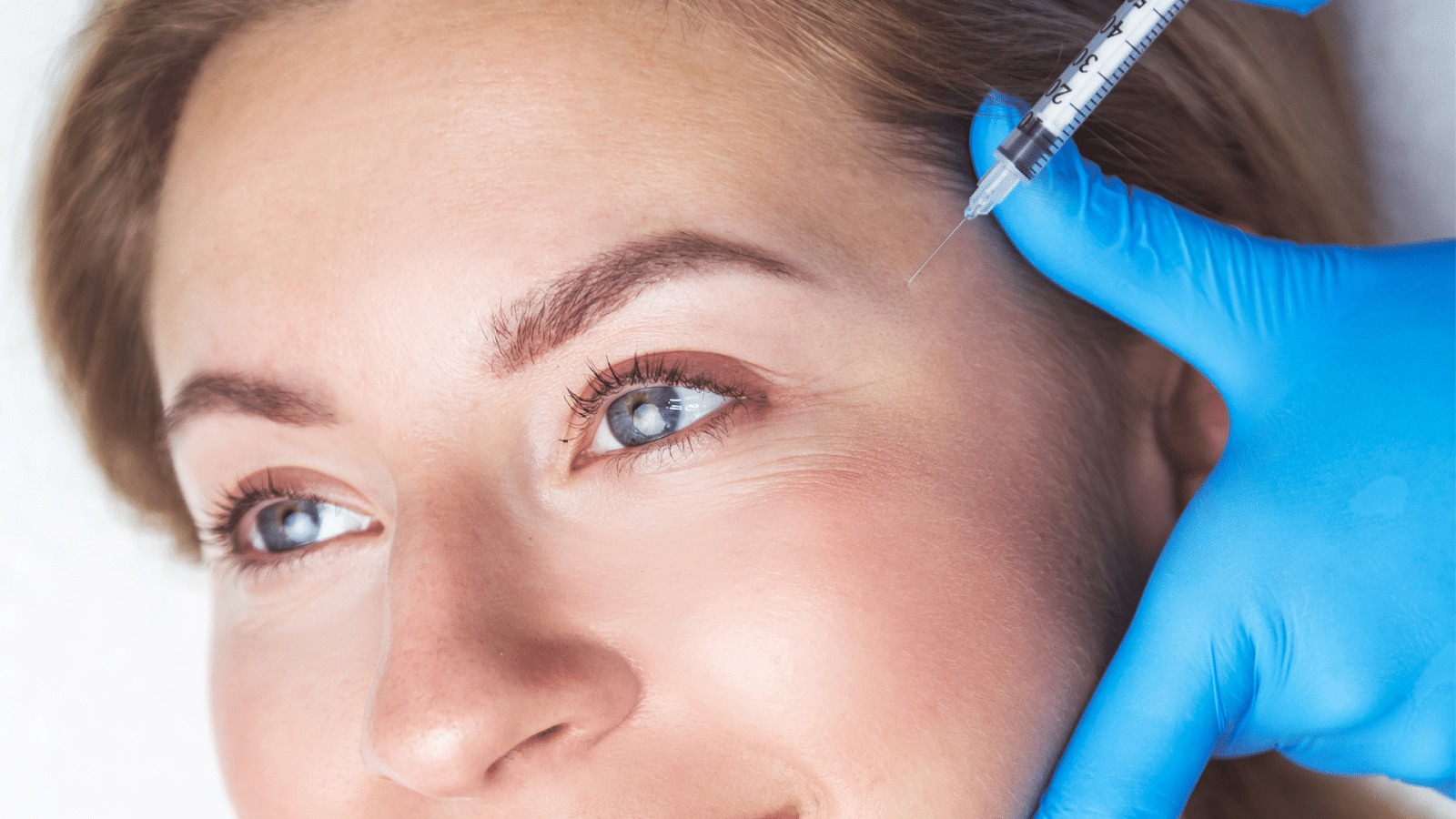 A woman with light hair is receiving an injection near her eye. Her face is relaxed, and she appears calm. A gloved hand holds the syringe carefully. The background is brightly lit.