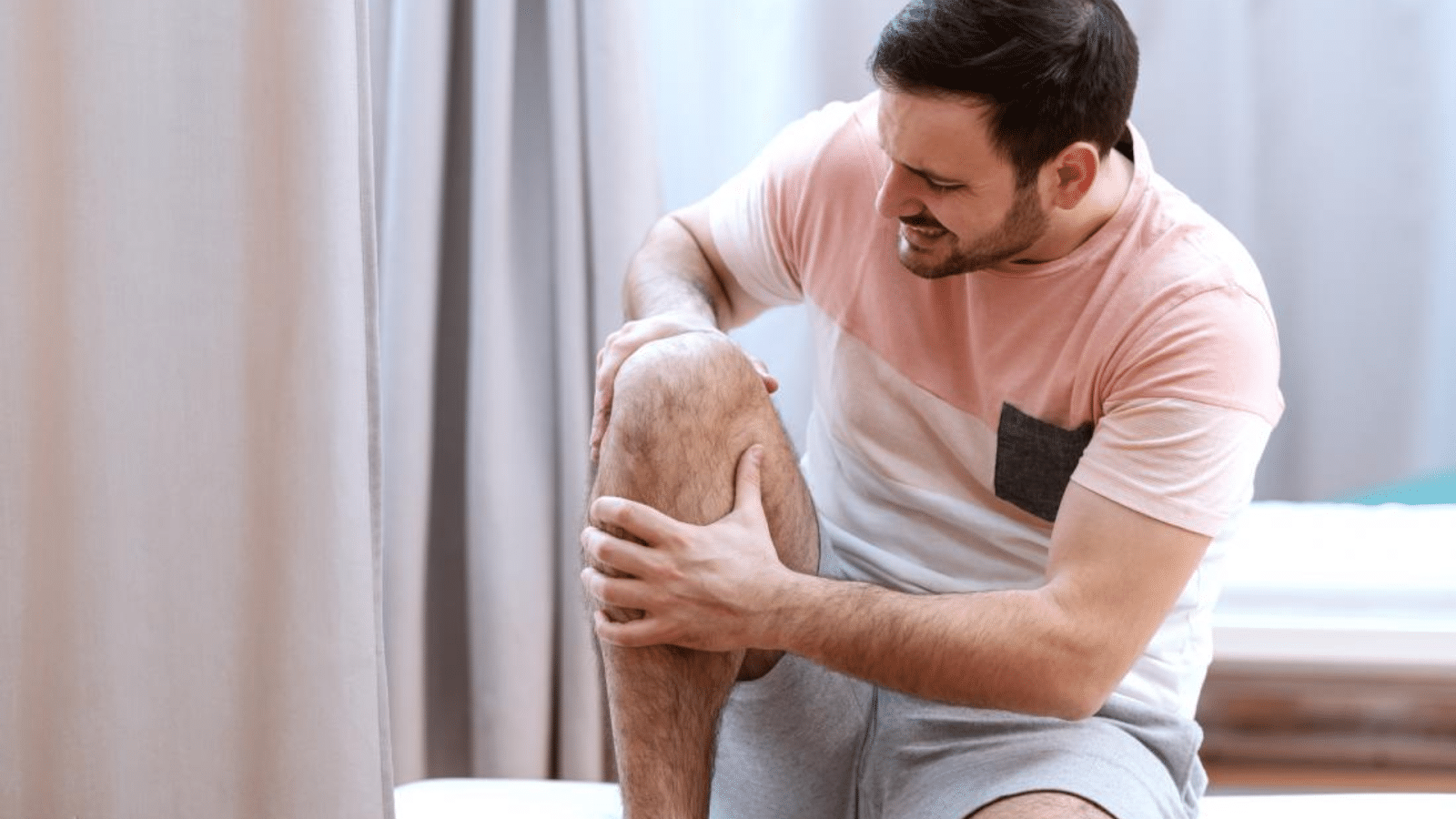 A man in a pink and gray shirt sits on a bed, holding his knee with both hands, and appears to be in pain. The background features beige curtains.