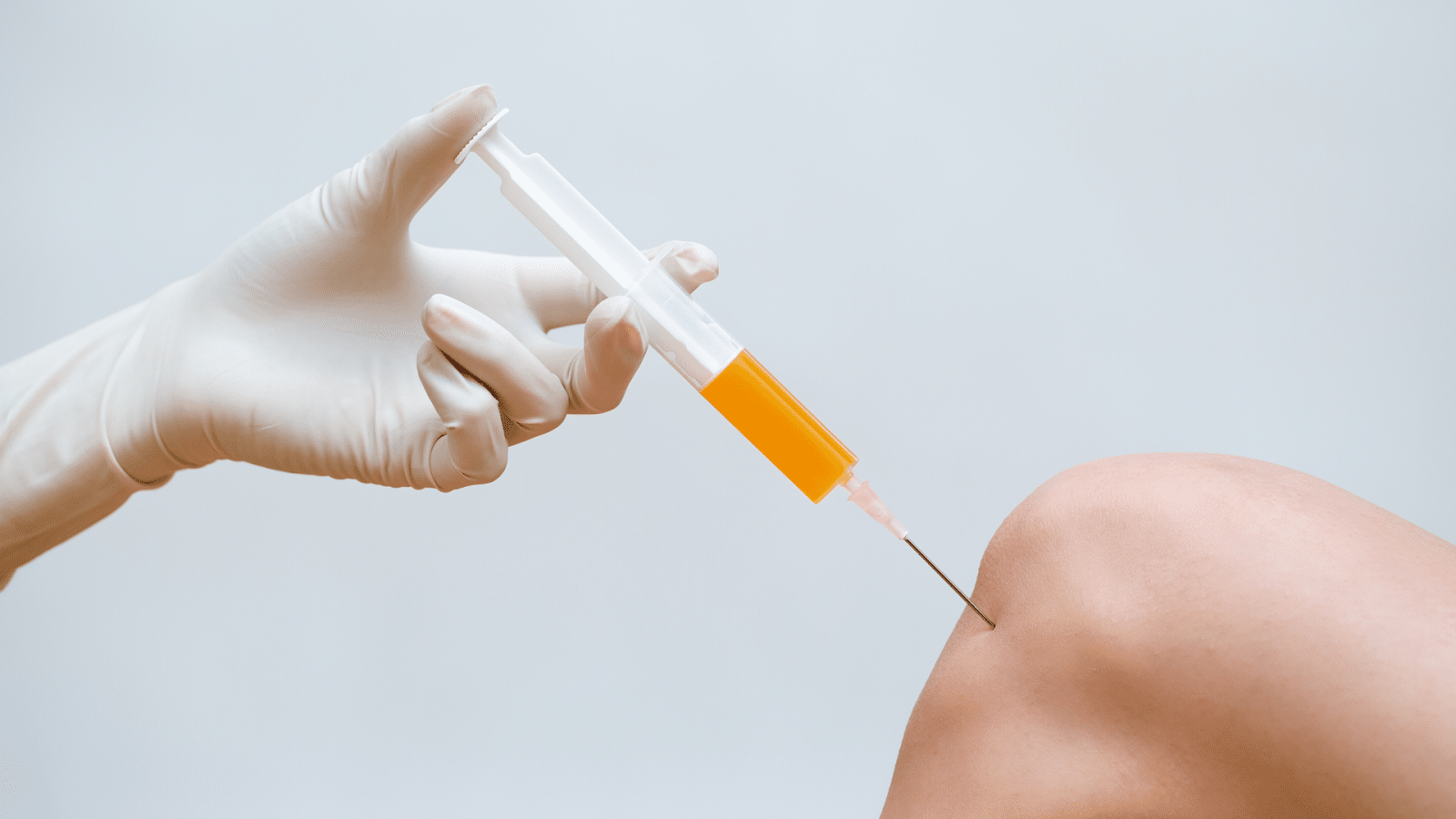 A gloved hand holds a syringe with yellow liquid, about to inject into a patient's knee against a plain background.