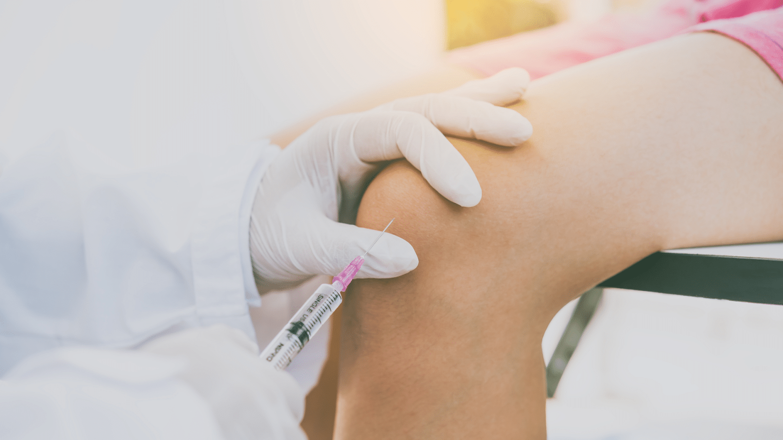 A medical professional wearing white gloves administers an injection to a patient's knee. The scene is well-lit, and the focus is on the careful procedure taking place.