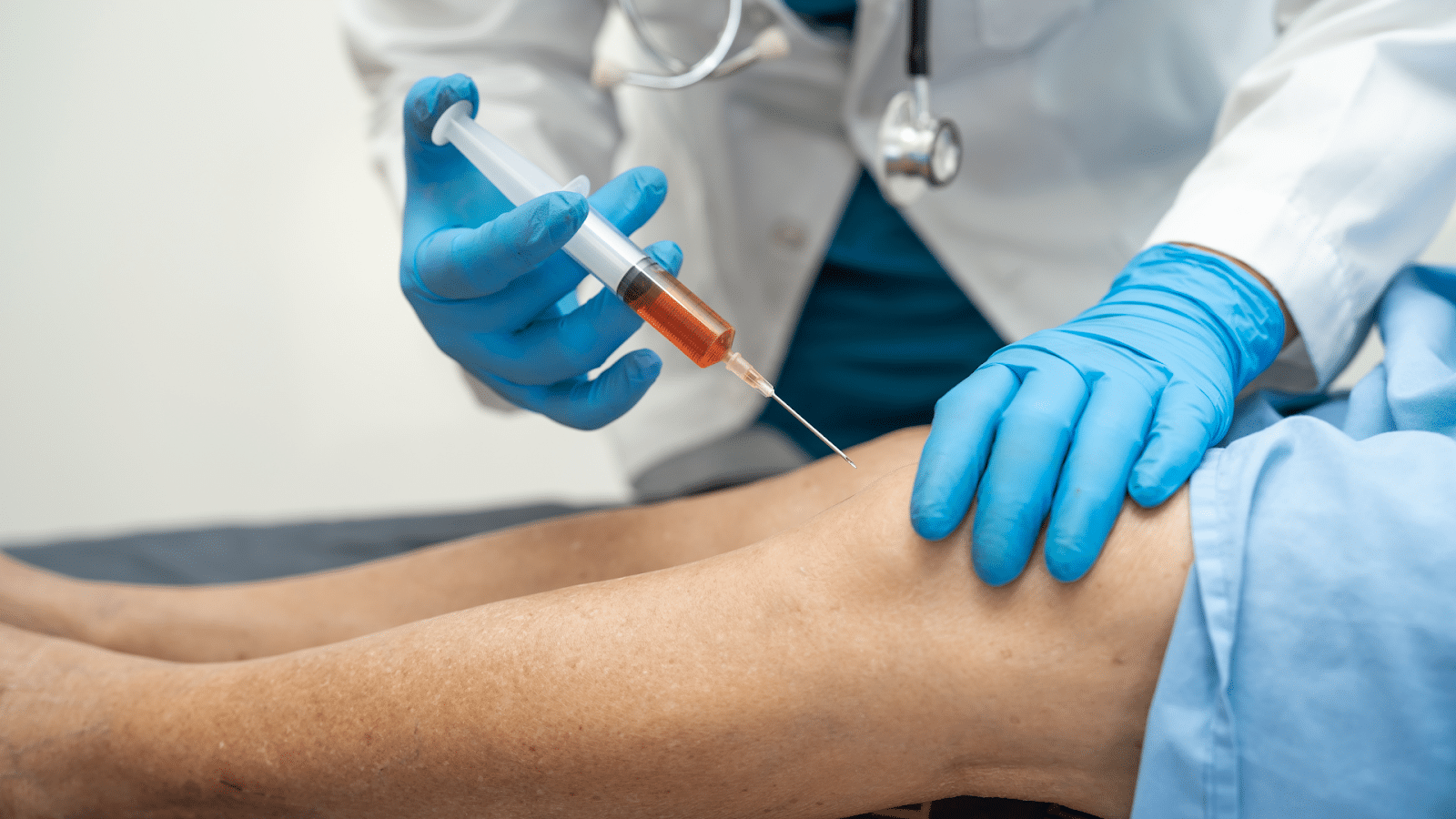 A healthcare professional wearing gloves injects a syringe into a patient's knee. The medical setting involves a person wearing a white coat, suggesting a clinical procedure or treatment.