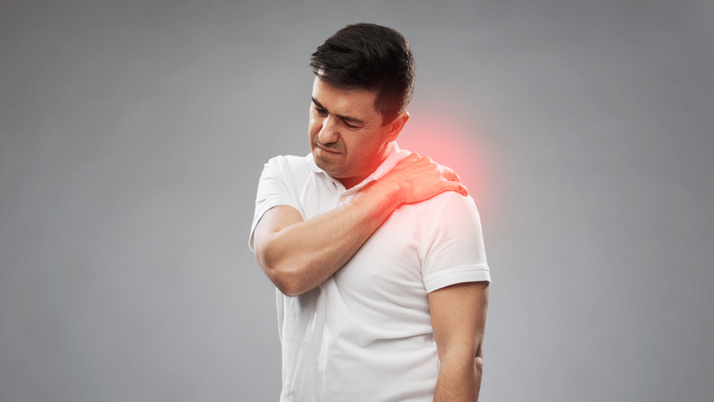 A man in a white shirt is holding his left shoulder, appearing to be in pain. The area he is touching is highlighted in red to indicate soreness or discomfort. The background is a plain grey.