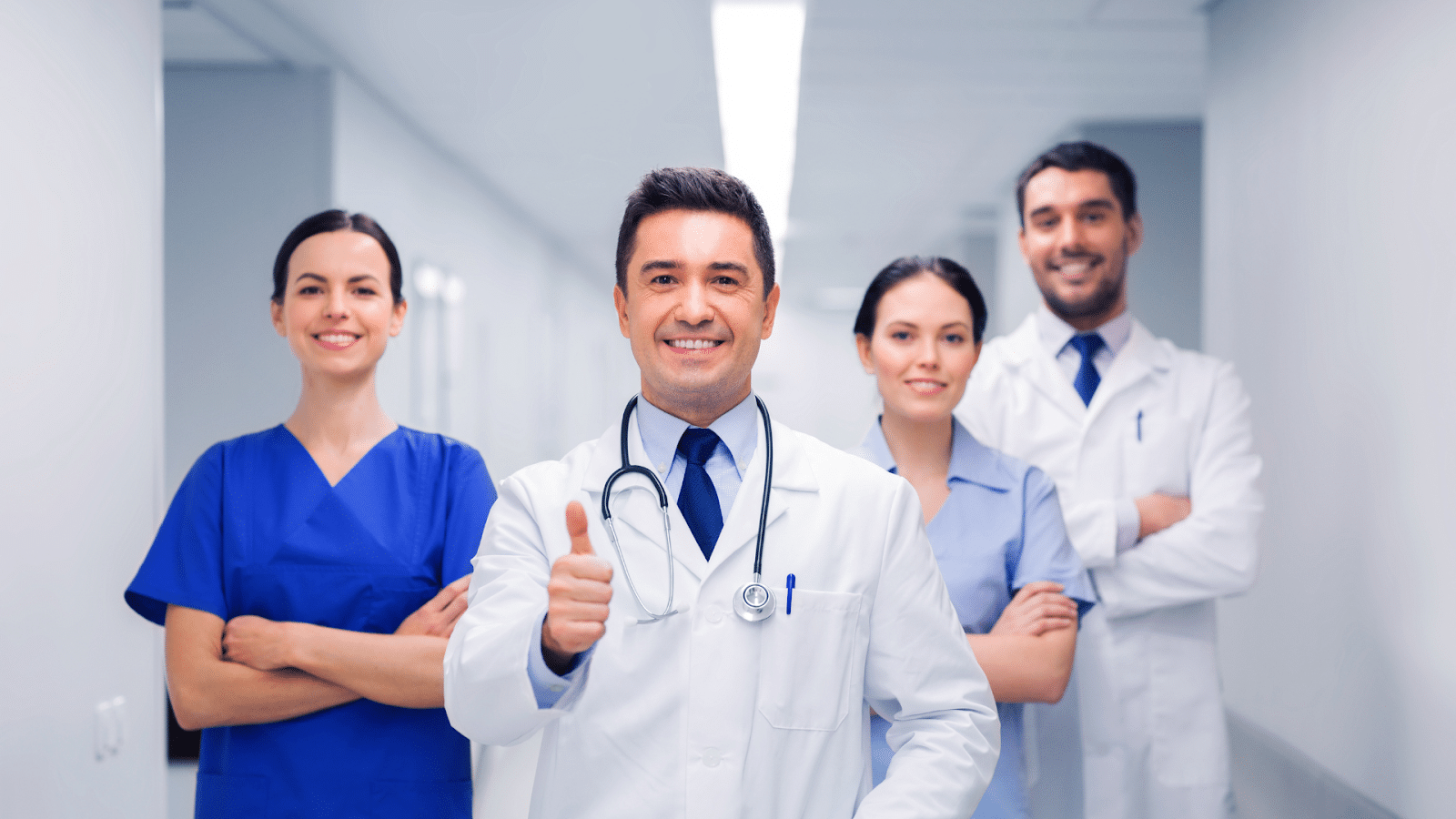 A group of four medical professionals in scrubs and lab coats stand in a hospital hallway. The man in front gives a thumbs-up, while the others stand with arms crossed, all smiling confidently.