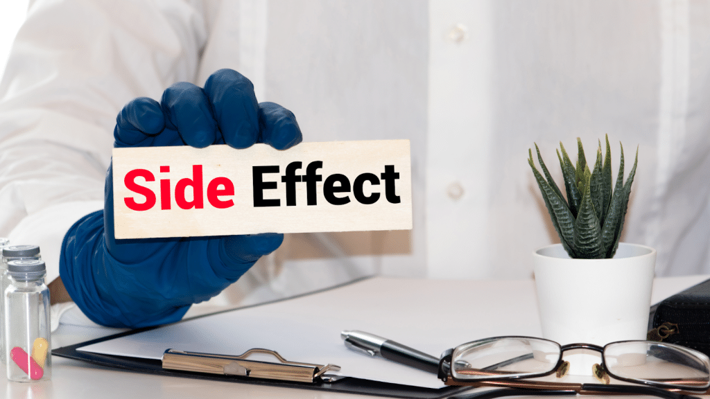 A person wearing a blue glove holds a sign that says "Side Effect" in red and black text. The scene includes pills, a small potted plant, a clipboard with papers, and a pair of glasses on a desk.