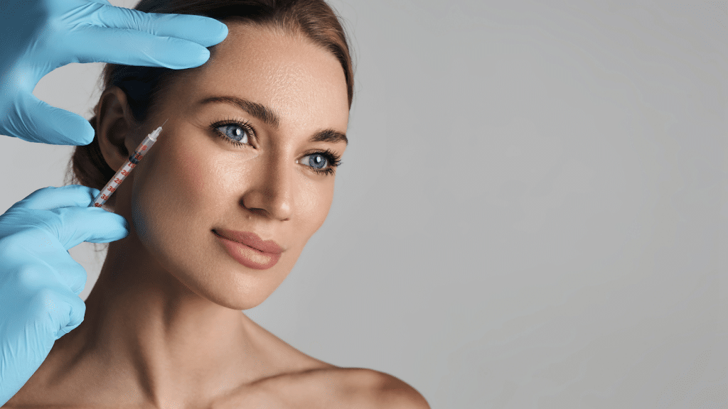 A person with blue eyes and light brown hair receives a cosmetic injection on the side of the face. They are surrounded by gloved hands, and the background is gray, highlighting the serene expression.