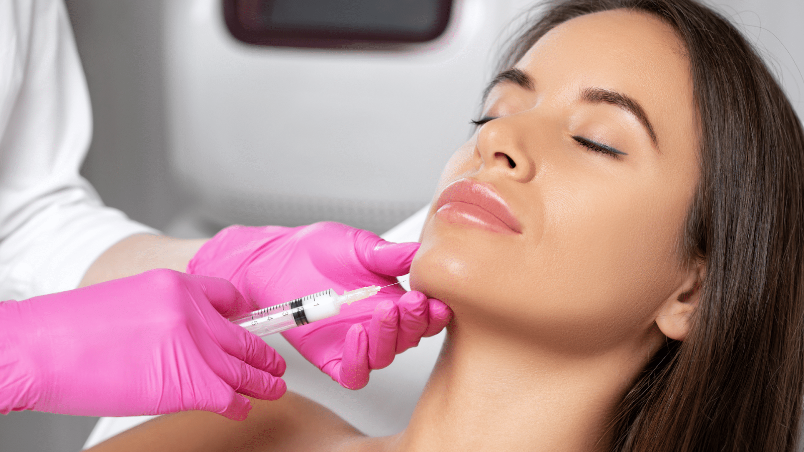 A woman with closed eyes is receiving an injection in the chin area. A person wearing pink gloves is administering the injection. The background is blurred, focusing on the procedure.