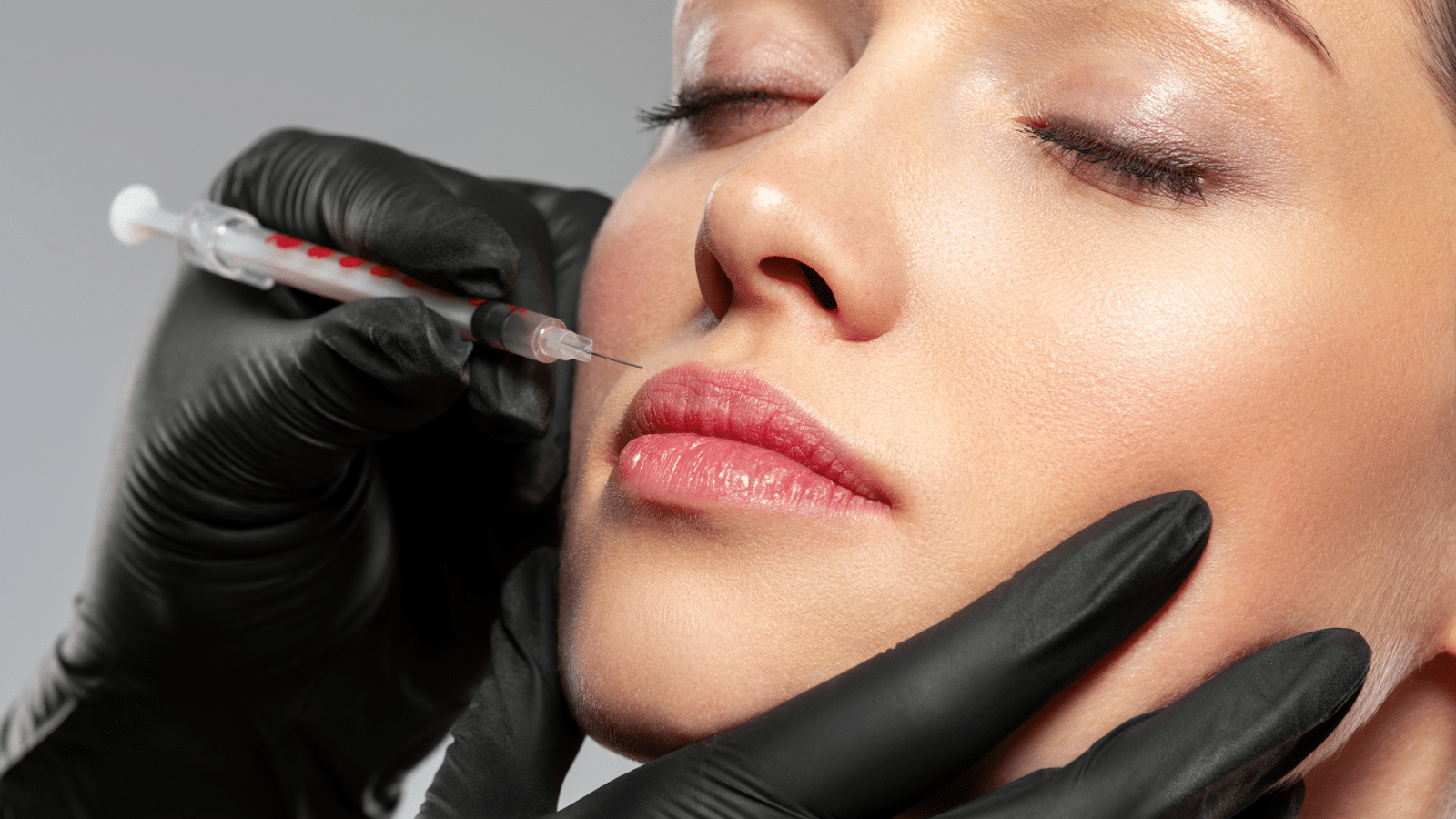 A close-up of a person receiving a cosmetic injection in the lips. The person has their eyes closed and is wearing neutral makeup. Gloved hands are holding a syringe close to the lip area against a neutral background.