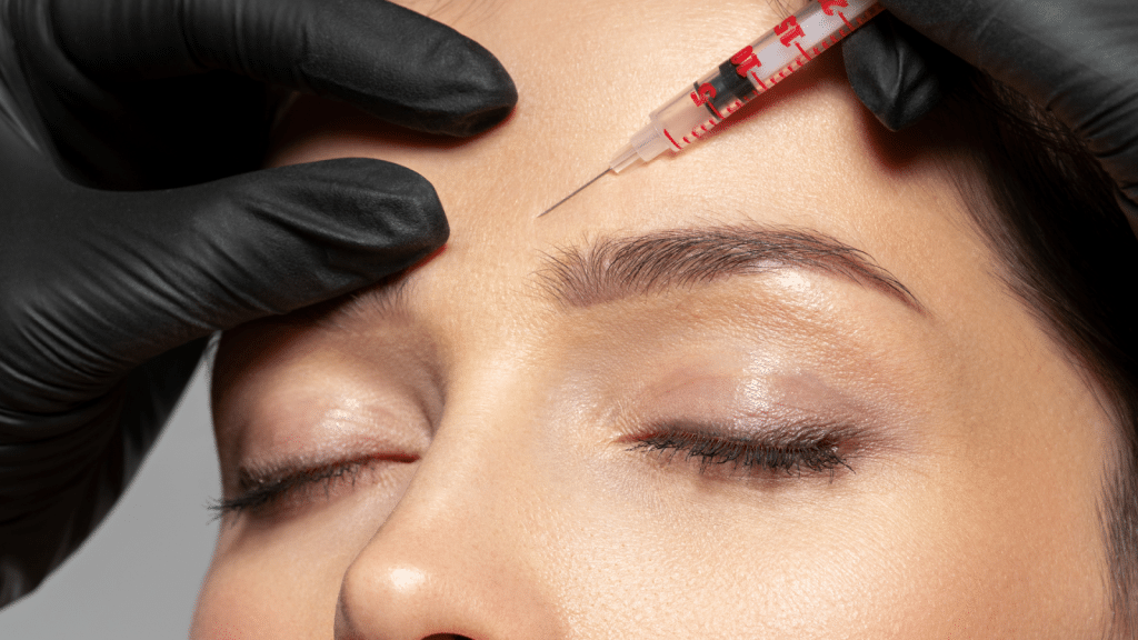 Close-up of a person with closed eyes, receiving an injection in the forehead. Hands in black gloves hold a syringe aimed at the brow area. The procedure appears to be a cosmetic treatment.