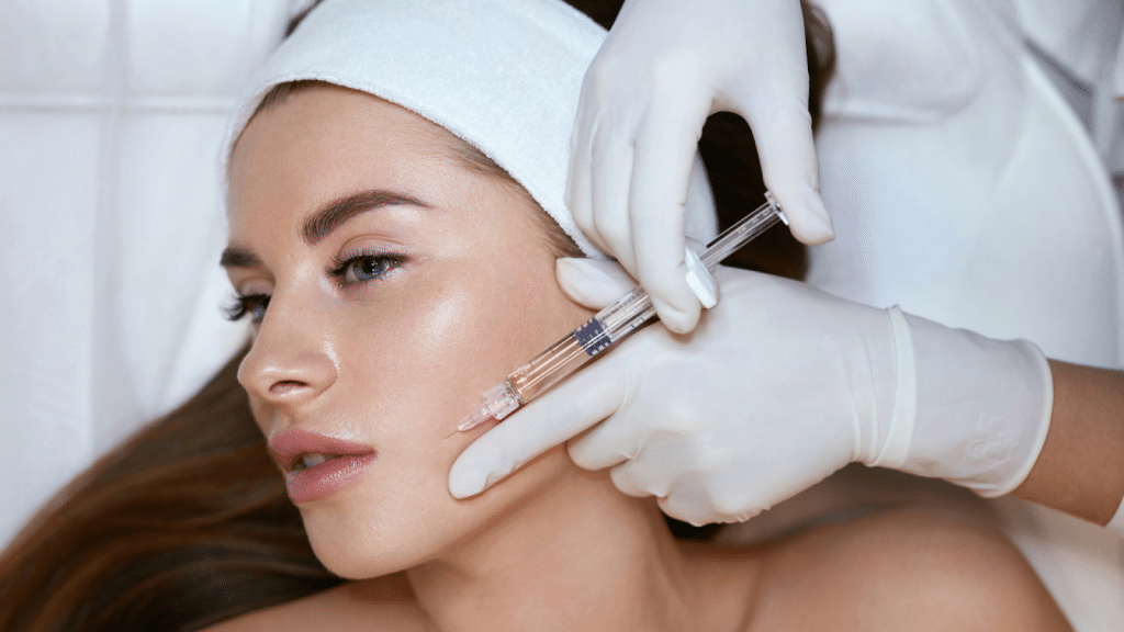 A person wearing a headband lies down while a gloved hand administers an injection to their cheek. The scene suggests a cosmetic or skincare procedure in a clinical setting.