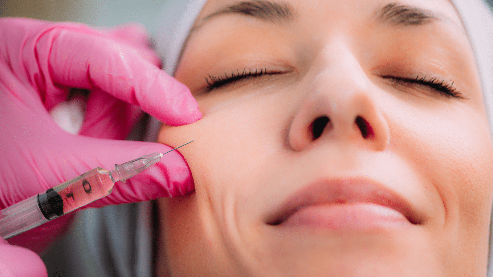 A person with closed eyes receives an injection on the cheek from a syringe held by a hand in pink gloves. The person’s face is relaxed, and they wear a white headband.