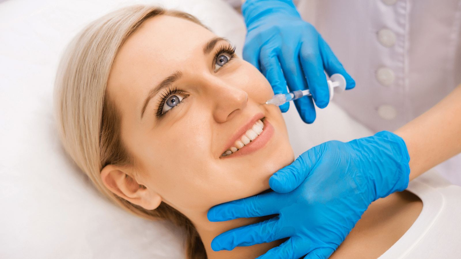 A woman with a serene expression lies on a treatment bed as a professional, wearing blue gloves, prepares to administer an injection on her face with a syringe.