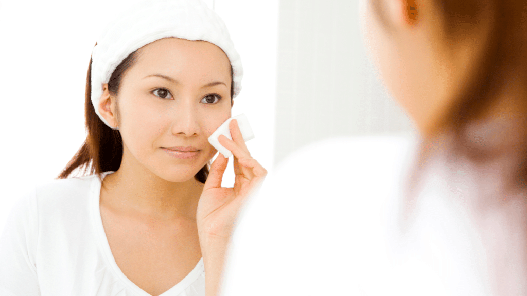 A woman with a headband is looking in the mirror while applying skincare. She gently uses a cotton pad on her cheek. The background is softly lit, creating a calm and clean atmosphere.