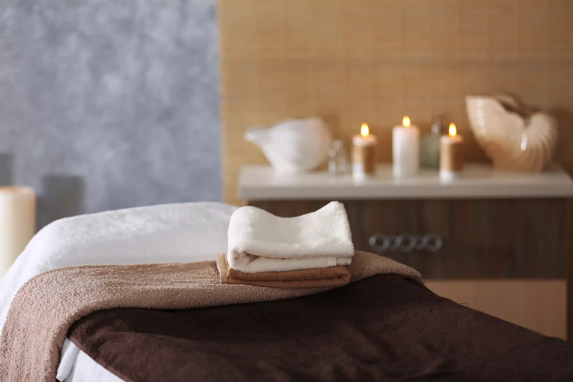 A cozy spa setting with soft towels folded neatly on a massage table. Candles are lit on a wooden console, casting a warm glow. The background features a textured wall and decorative elements, creating a calming atmosphere.