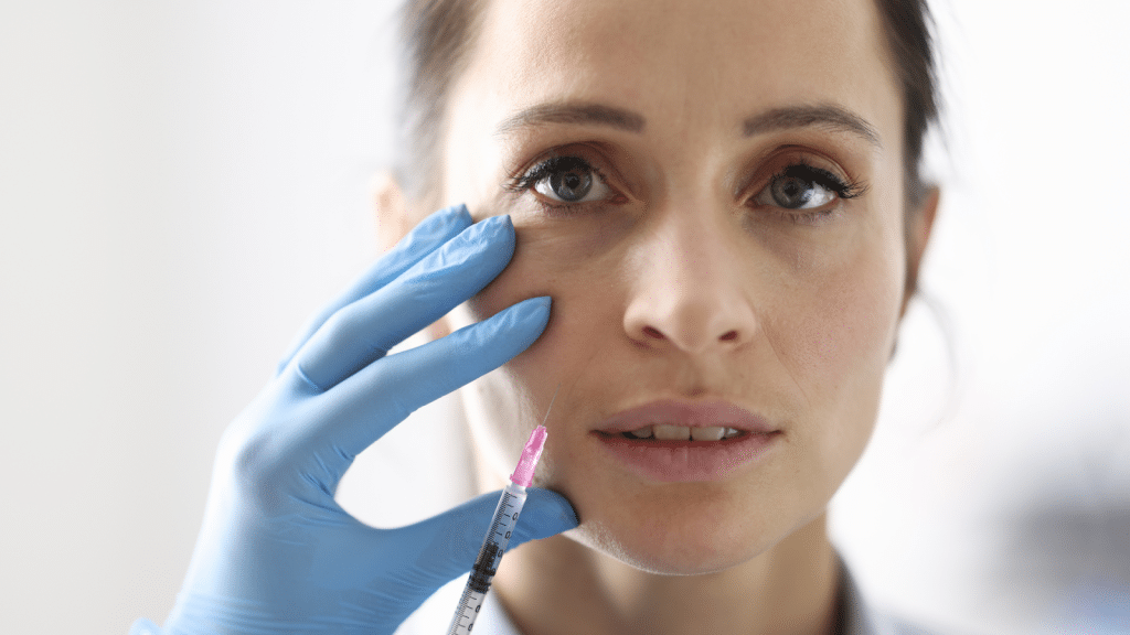 A woman with blue gloves is holding a syringe near her cheek, preparing for an injection. She has light skin and brown hair, and her expression is neutral. The background is blurred and white, suggesting a clinical setting.