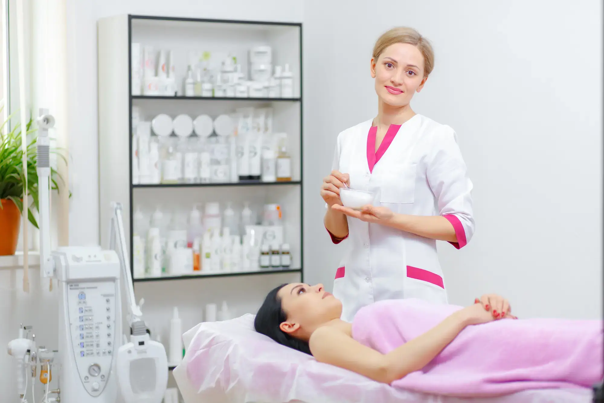 A beautician in a white and pink uniform stands smiling, holding a bowl, next to a client lying on a treatment bed covered with a pink towel. The room is bright, with shelves of beauty products in the background.