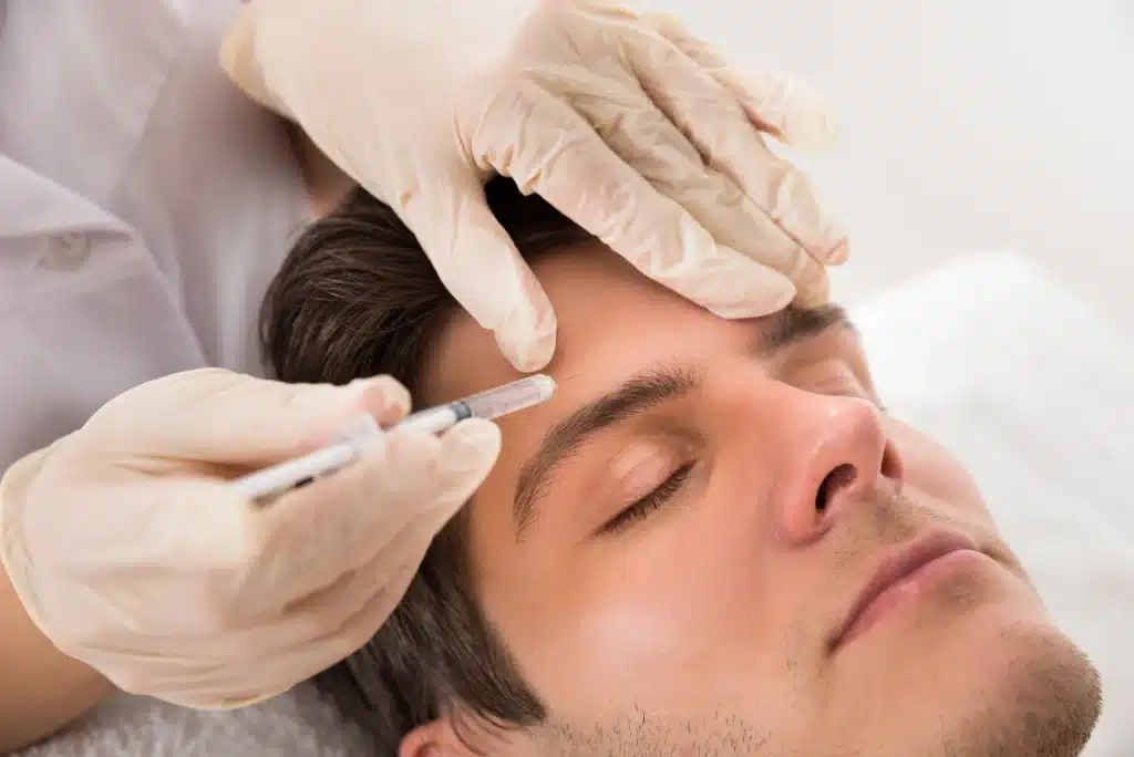 A person with closed eyes lying down receives an injection in the forehead. A practitioner wearing gloves carefully administers the procedure in a clinical setting.