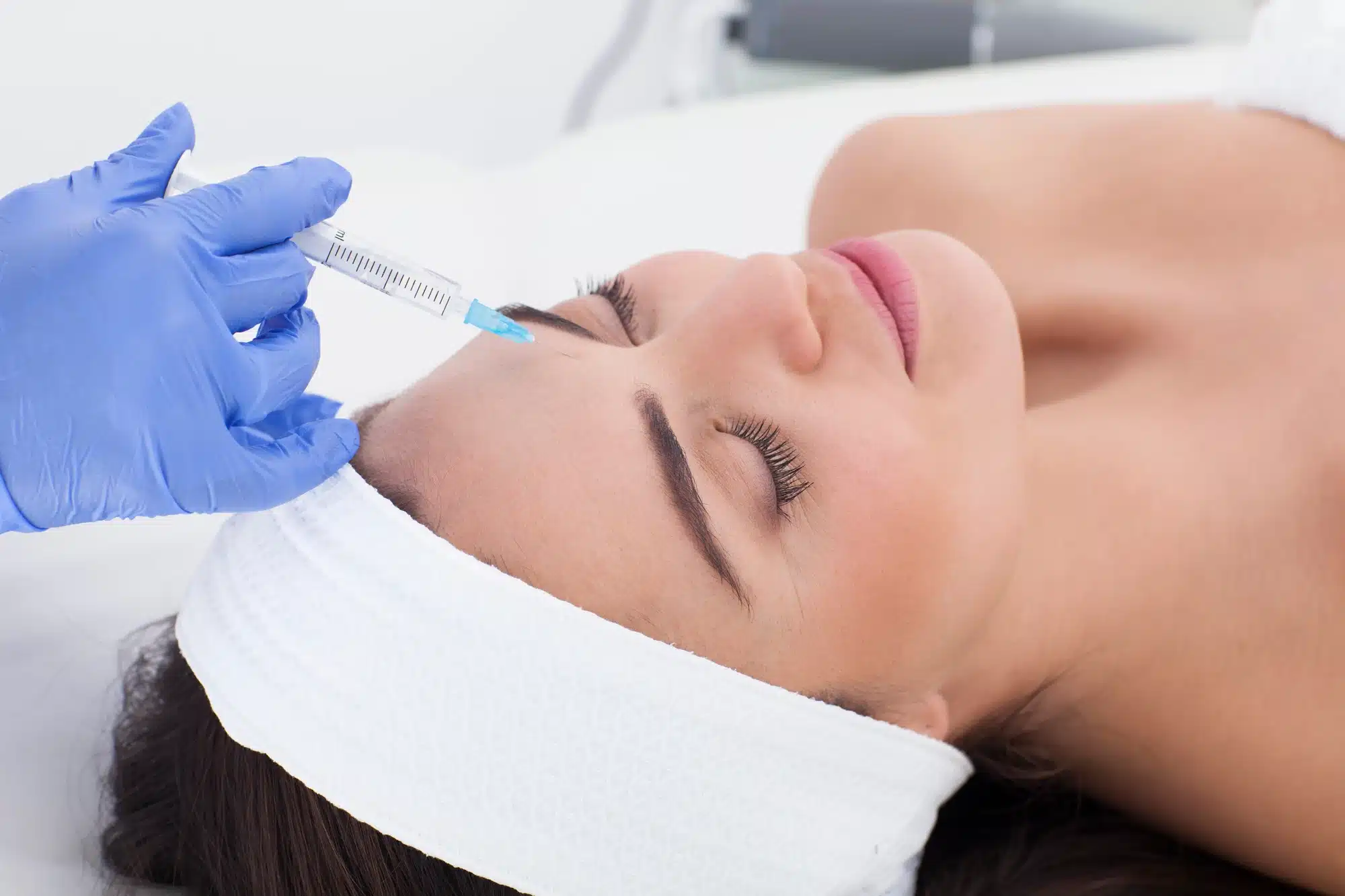 A person with a headband is lying on a bed, receiving an injection on their forehead from a gloved hand holding a syringe. The setting appears to be a clinical or cosmetic procedure environment.